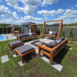 WOJCIECH GRĘBOWICZ - Drewniany zestaw mebli ogrodowych z poduchami, w tym huśtawka, otaczający palenisko na trawniku, w tle basen i zabudowa. Słoneczny dzień.