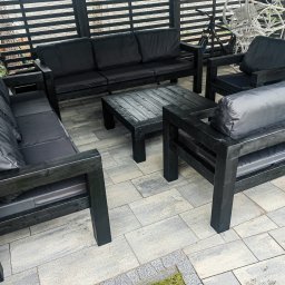 WOJCIECH GRĘBOWICZ - Komplet mebli ogrodowych z ciemnego drewna pod zadaszeniem. Na drugim planie widoczny rower i wiszące krzesło. Całość na tle ściany z kamienia.