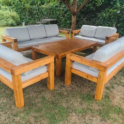 WOJCIECH GRĘBOWICZ - Drewniany komplet mebli ogrodowych: stół i sofy z jasnymi poduchami, ustawione na trawie pod zadaszeniem. Naturalne drewno, solidna konstrukcja.