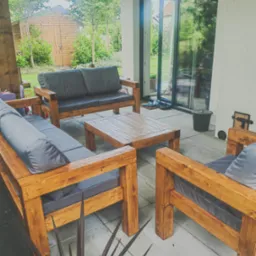 Drewniany komplet wypoczynkowy (sofa, fotele, stół) z szarymi poduchami na tarasie z widokiem na ogród i drewnianą szopę. Widoczne przesuwne drzwi tarasowe.