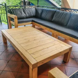 Drewniany stół i narożnik z czarnymi poduchami na tarasie z kutą balustradą. Naturalne drewno i ciemne poduszki tworzą kontrast na tle brązowej podłogi.