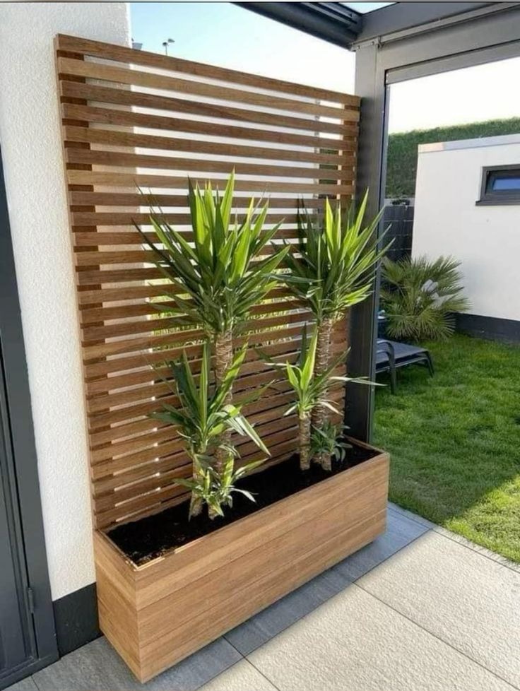 Drewniana konstrukcja z poziomych listew, pełniąca funkcję osłony i podpory dla roślin w podłużnej donicy, ustawiona na tarasie przy ścianie budynku.