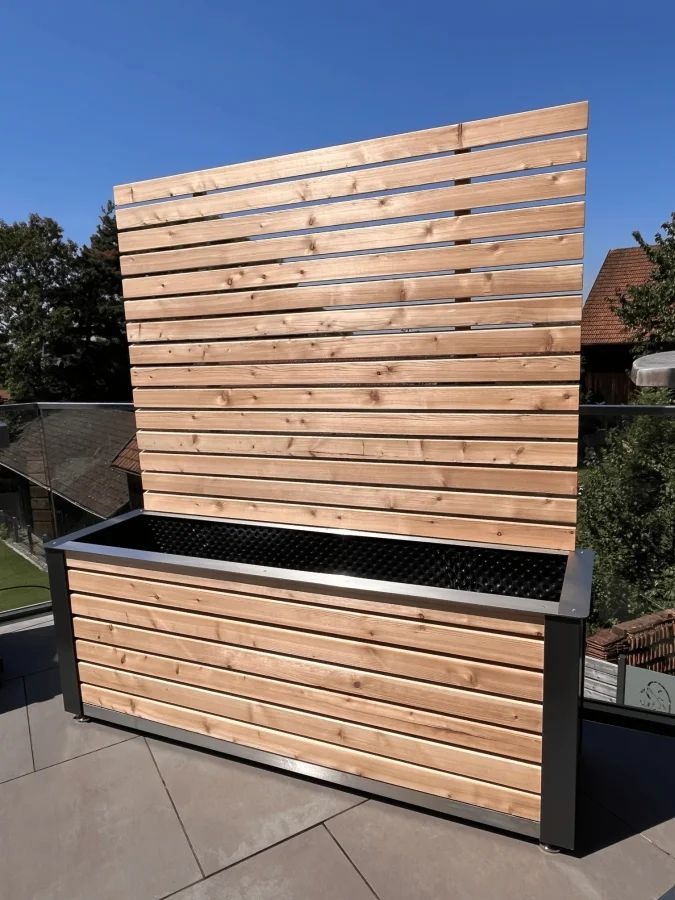 Nowoczesna drewniana donica z panelem osłonowym, wykonana z jasnego drewna z metalowymi akcentami, na tarasie z widokiem na dom i błękitne niebo.