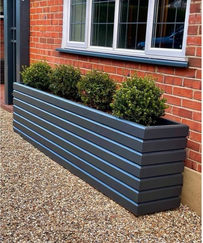 Długa, prostokątna, ciemnoszara donica z poziomych desek z jasnymi przerwami, ustawiona przed ceglaną ścianą domu i oknem, wypełniona trzema kulistymi krzewami na żwirowym podłożu.