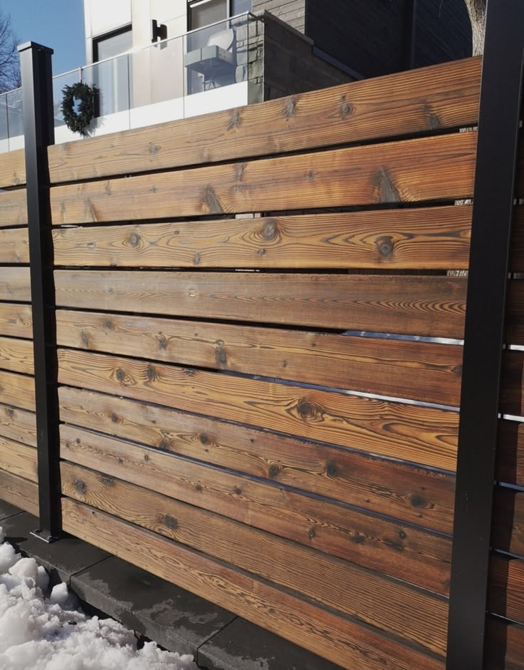 Nowoczesne ogrodzenie z poziomo ułożonych, impregnowanych desek z widocznym usłojeniem, czarne metalowe słupki, śnieg u podstawy, budynek w tle.