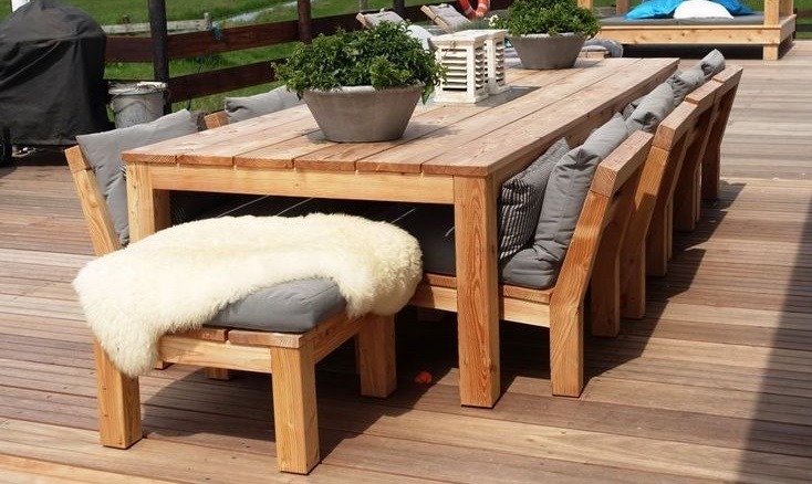 Duży, drewniany stół z ławkami i krzesłami na drewnianym tarasie, z szarymi poduszkami i donicami z zielonymi roślinami, obok grill i leżanka.