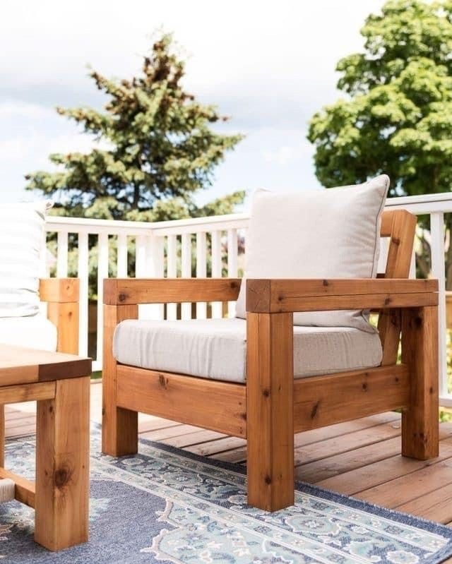 Rustykalny fotel ogrodowy z jasnego drewna na tarasie, z poduszką w kolorze ecru i dywanem w odcieniach błękitu, widok na drzewa i białą balustradę.