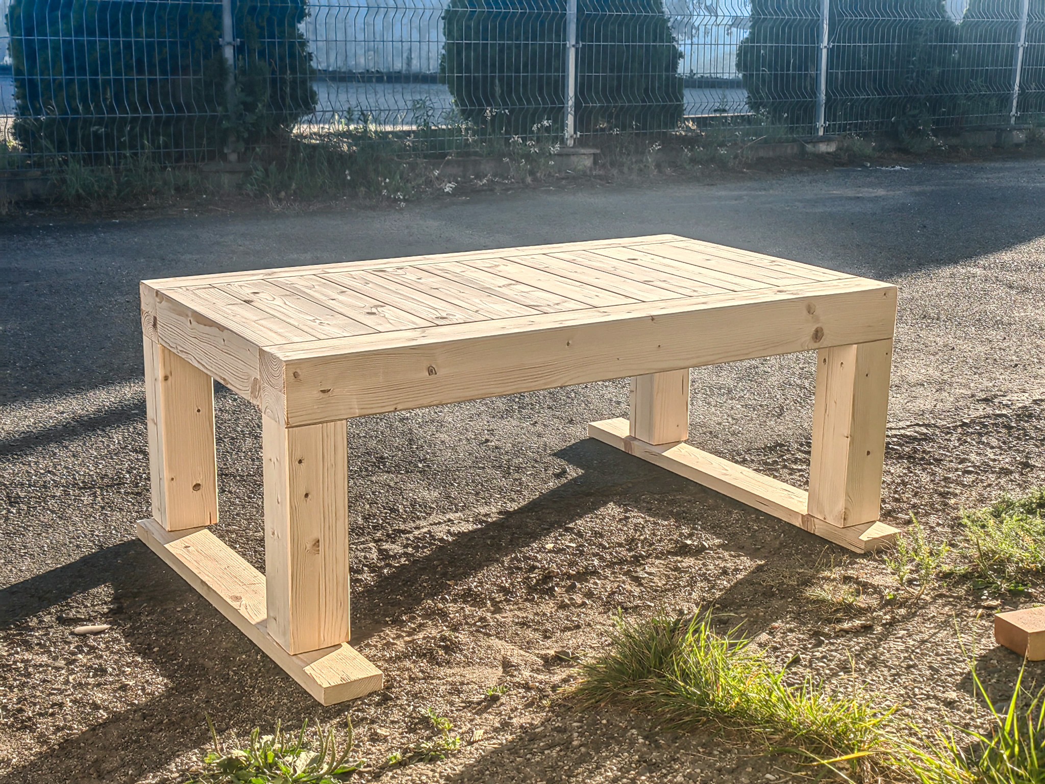 Surowy drewniany stół ogrodowy z litego drewna na zewnątrz, z widocznymi słojami, w słoneczny dzień. Konstrukcja prosta, minimalistyczna, bez wykończenia.