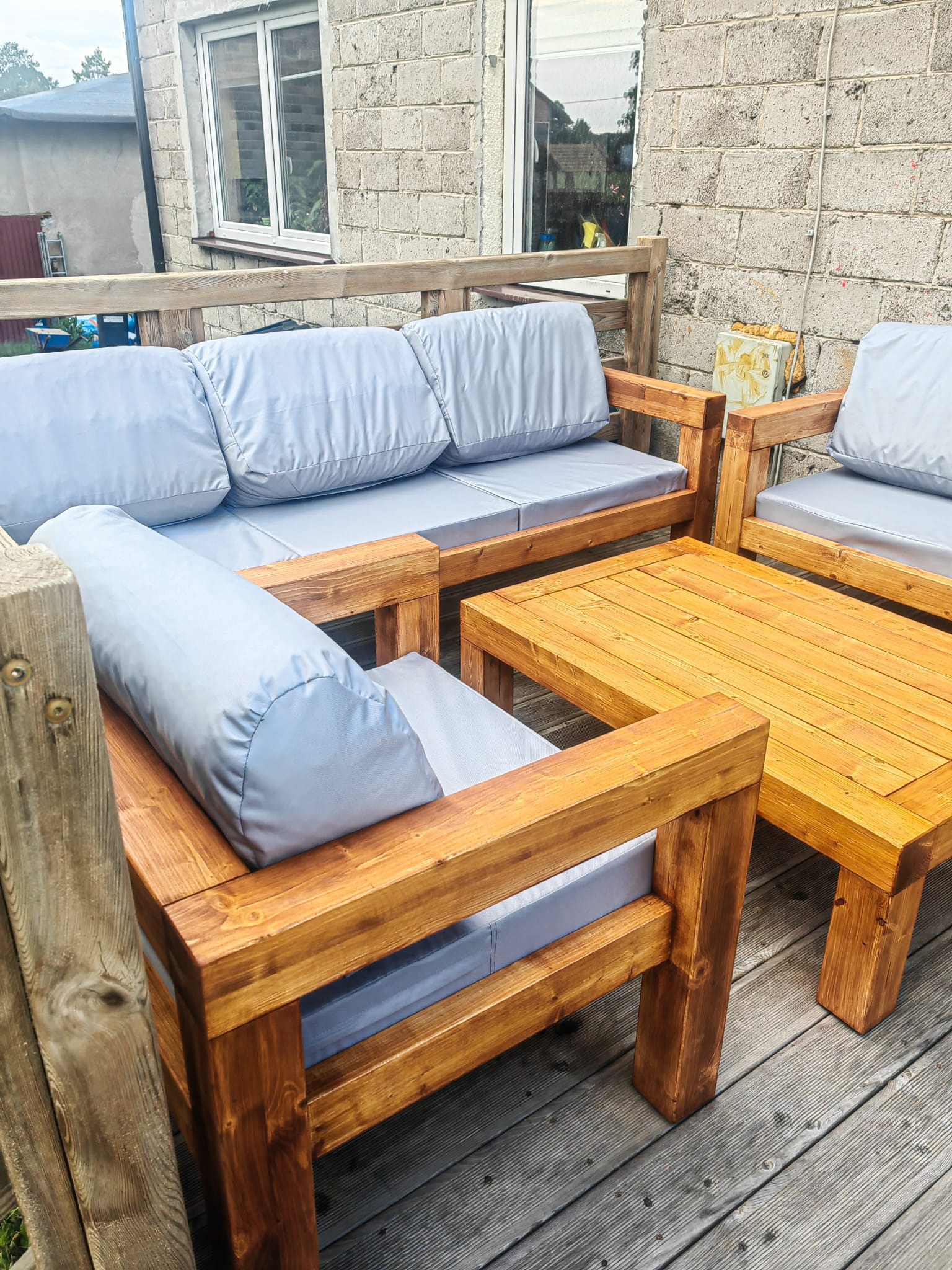 Komplet drewnianych mebli ogrodowych z jasnymi poduchami na tarasie, widok z bliska, z ciemnobrązowym wykończeniem drewna i szarymi deskami podłogowymi.