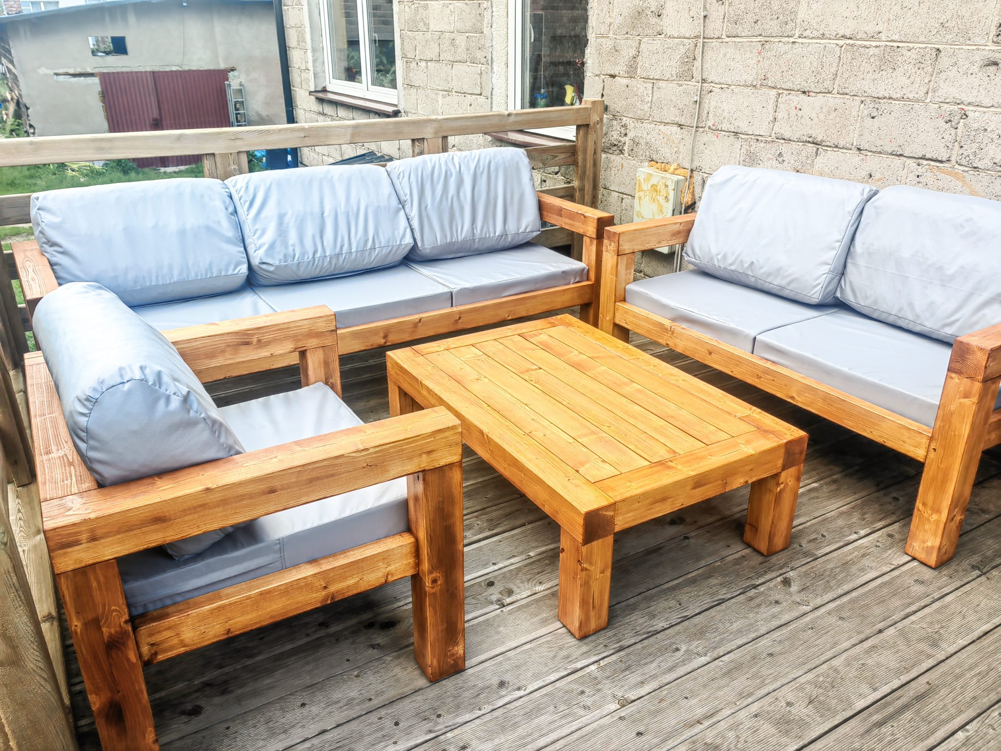 Zestaw mebli ogrodowych z jasnego drewna i szarymi poduszkami na tarasie z desek. Widoczna sofa, dwa fotele i stolik kawowy. Tło stanowi ściana z bloczków.