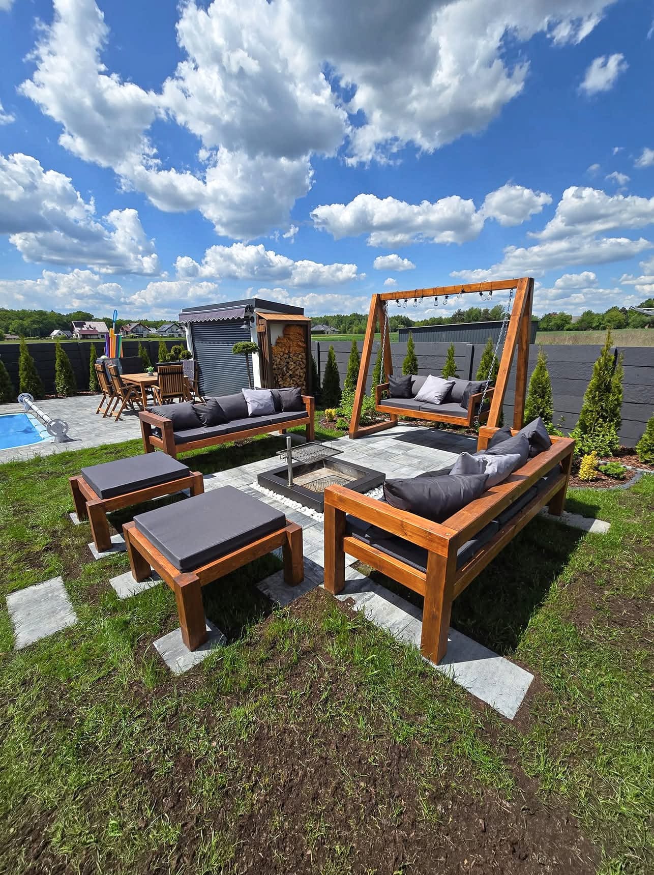Drewniany zestaw mebli ogrodowych z poduchami, w tym huśtawka, otaczający palenisko na trawniku, w tle basen i zabudowa. Słoneczny dzień.