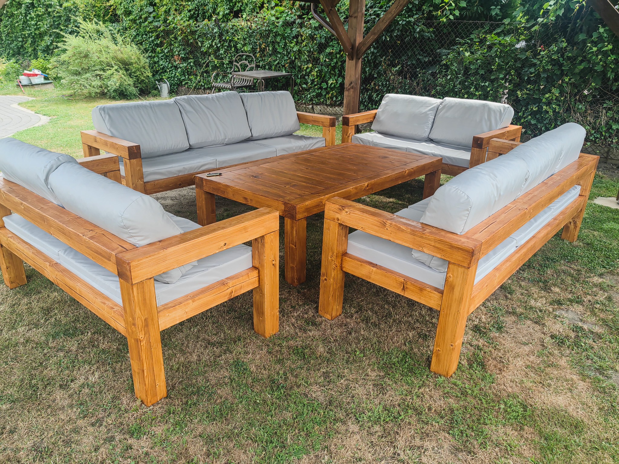 Drewniany komplet mebli ogrodowych: stół i sofy z jasnymi poduchami, ustawione na trawie pod zadaszeniem. Naturalne drewno, solidna konstrukcja.