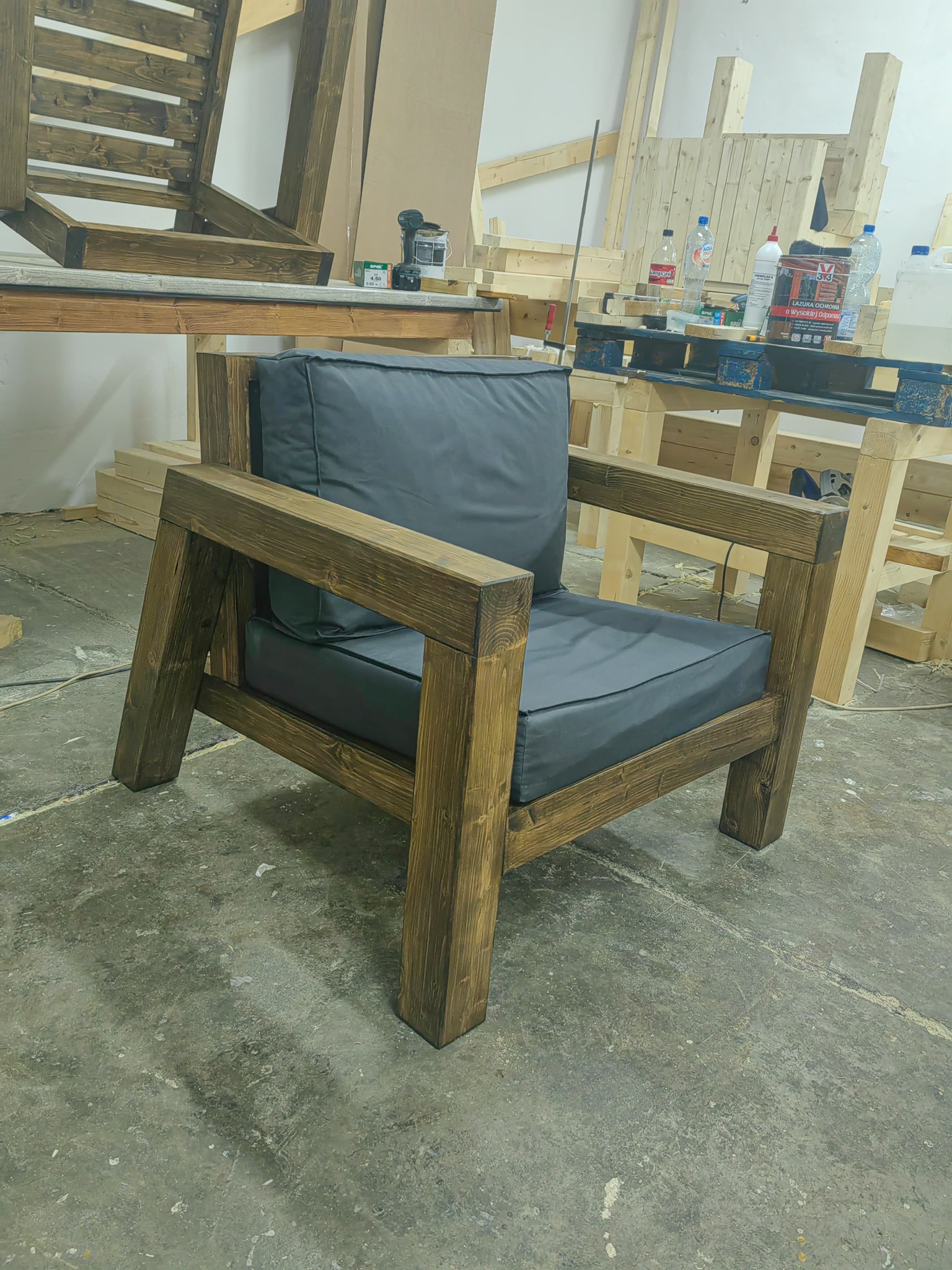 Rustykalny fotel z ciemnego drewna z szarymi poduszkami, wykonany ręcznie. W tle warsztat stolarski z narzędziami i elementami konstrukcyjnymi z drewna.