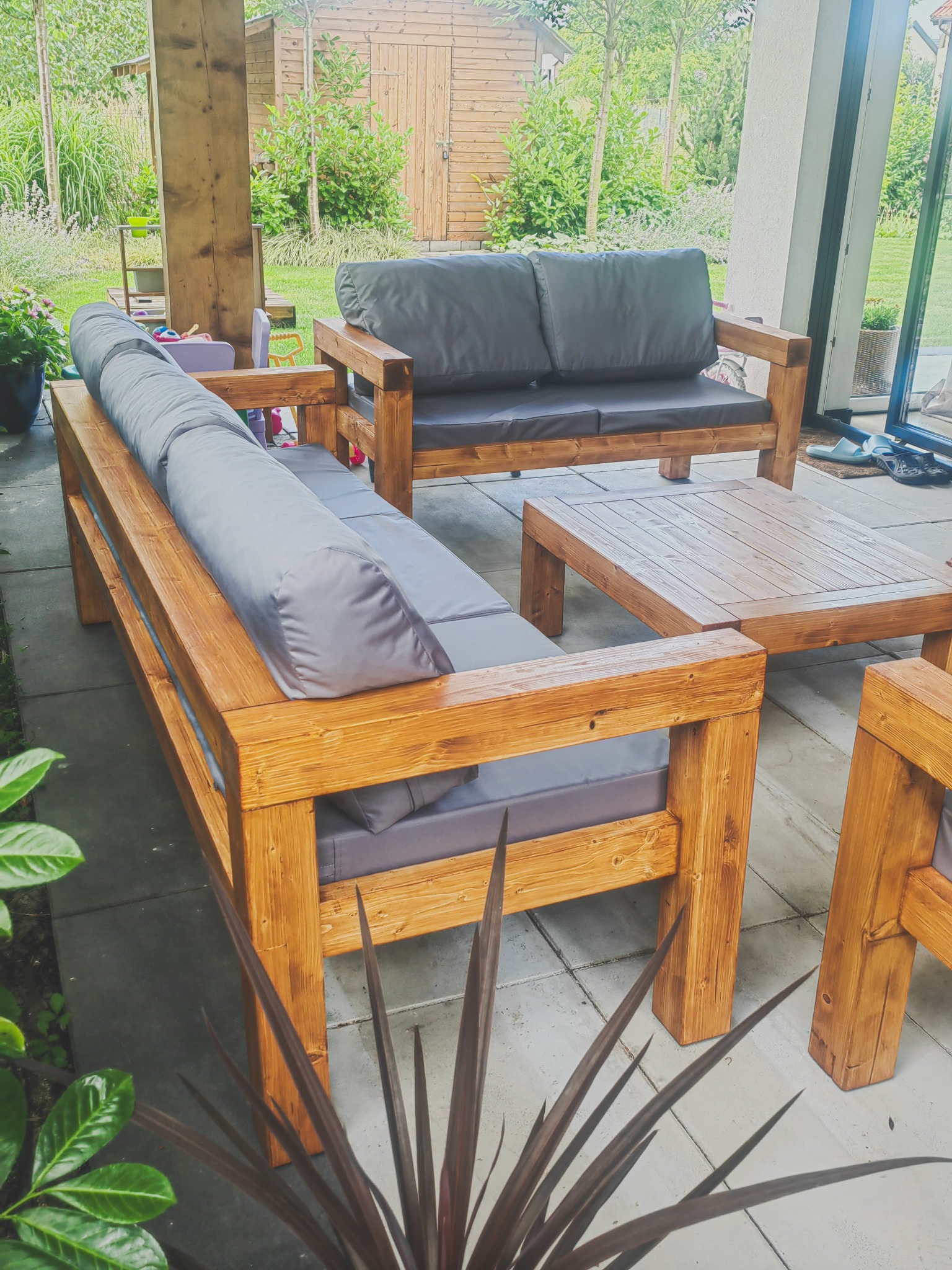 Rustykalny zestaw mebli ogrodowych z drewna: sofa z poduchami, fotel i stolik kawowy, na tle zielonego ogrodu i drewnianej altany. Widoczne detale wykończenia drewna.