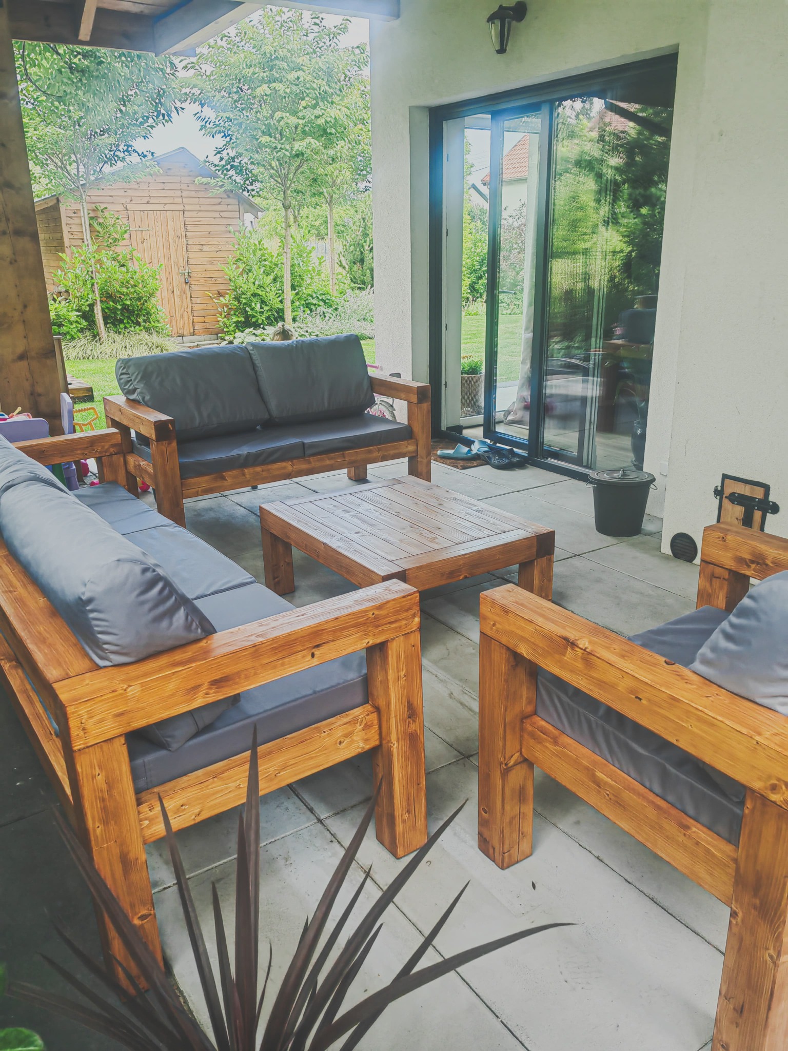 Drewniany komplet wypoczynkowy (sofa, fotele, stół) z szarymi poduchami na tarasie z widokiem na ogród i drewnianą szopę. Widoczne przesuwne drzwi tarasowe.