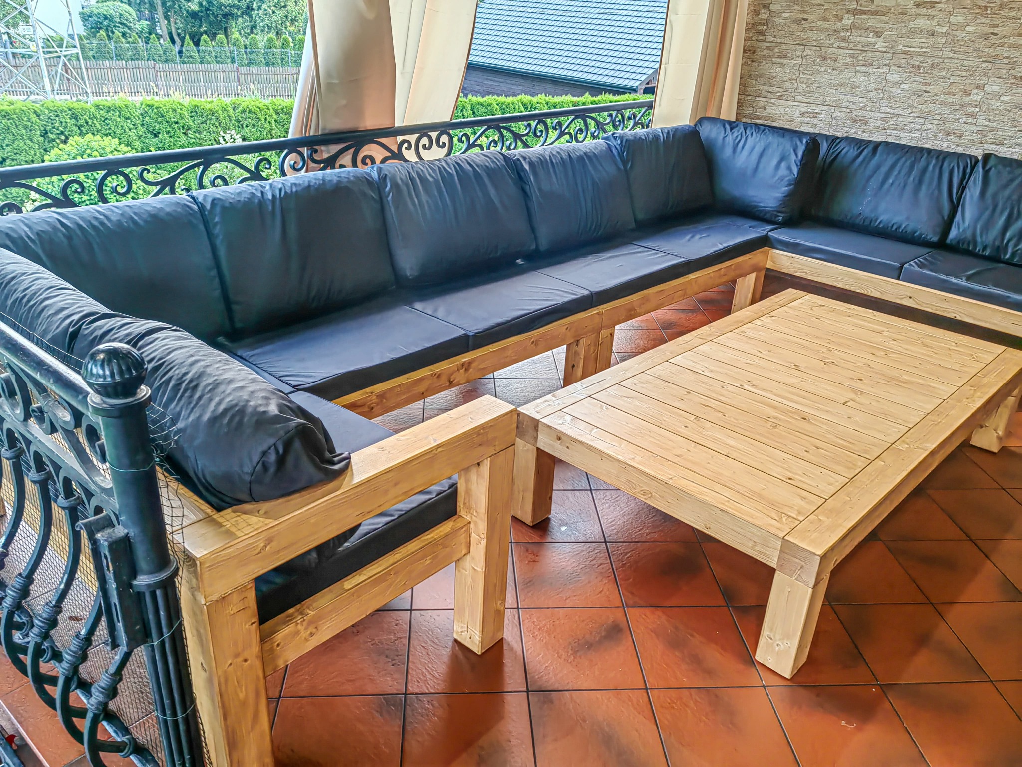 Kącik wypoczynkowy z drewnianym stołem i narożnikiem z czarnymi poduchami na tarasie z kutą balustradą i widokiem na zieleń. Ciepłe światło podkreśla fakturę płytek.