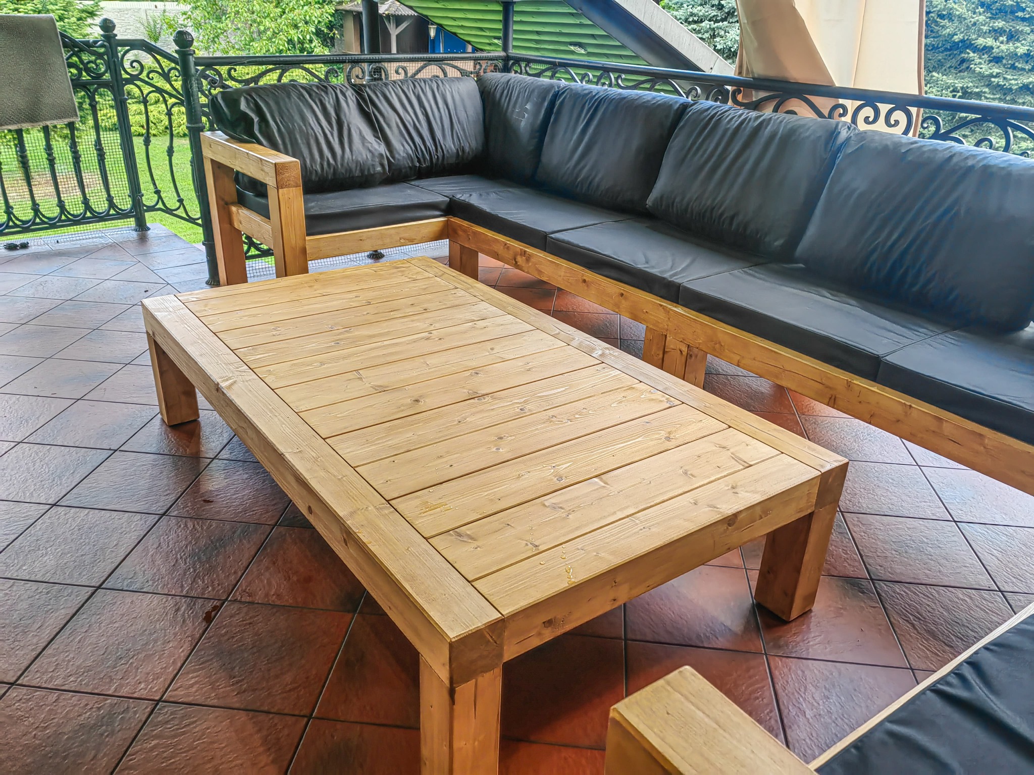 Drewniany stół i narożnik z czarnymi poduchami na tarasie z kutą balustradą. Naturalne drewno i ciemne poduszki tworzą kontrast na tle brązowej podłogi.