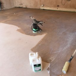 TOM INSTAL - BUD - Gruntowanie podłogi w pomieszczeniu: widoczne dwa białe kanistry z etykietą Ultra Floor Prime II Primer, wałek malarski z długim uchwytem oraz pompa z przewodami, podłoga częściowo pokryta...