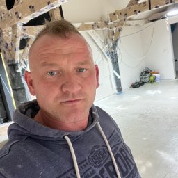 TOM INSTAL - BUD - Mężczyzna w bluzie z kapturem robi selfie na tle remontowanego pomieszczenia z zabezpieczonymi folią belkami stropowymi i narzędziami w tle.