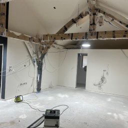 TOM INSTAL - BUD - Wnętrze w trakcie remontu: zabezpieczone folią belki stropowe, świeżo pomalowane ściany, wystające przewody elektryczne, sprzęt budowlany na posadzce.