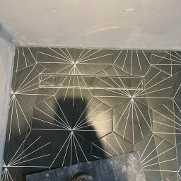 TOM INSTAL - BUD - Czarno-biała, geometryczna posadzka w łazience z odpływem liniowym, częściowo zakryta brudną deską i butem roboczym.