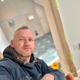 TOM INSTAL - BUD - Autoportret mężczyzny w bluzie z kapturem na tle remontowanej łazienki z widoczną podłogą w geometryczne wzory i zabezpieczonymi kartonem framugami drzwi.