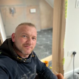 TOM INSTAL - BUD - Autoportret rzemieślnika w bluzie roboczej z plamami farby, na pierwszym planie narzędzia, w tle świeżo ułożona podłoga z geometrycznym wzorem w szarościach i fragment ściany w trakcie prac...