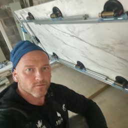 TOM INSTAL - BUD - Glazurnik w niebieskiej czapce, robiący selfie na tle płyty imitującej marmur, przymocowanej przyssawkami do ramy podczas transportu lub montażu.