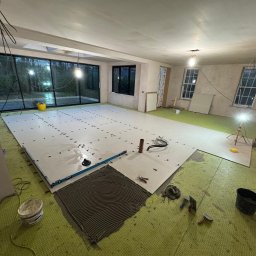 TOM INSTAL - BUD - Wnętrze w trakcie układania jasnych, wielkoformatowych płytek podłogowych z użyciem poziomicy i krzyżyków dystansowych; widoczny fragment nałożonego kleju i narzędzia glazurnicze.