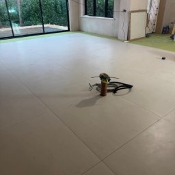 TOM INSTAL - BUD - Jasna, świeżo położona podłoga z dużych, kwadratowych płytek w surowym wnętrzu z dużymi oknami, na środku widoczny pionowy element z narzędziami i kablami.