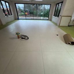 TOM INSTAL - BUD - Ułożona jasna podłoga z płytek w przestronnym pomieszczeniu z dużymi oknami z widokiem na zewnątrz, narzędzia i rolka papieru ochronnego na podłodze.