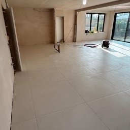 TOM INSTAL - BUD - Wnętrze w trakcie remontu z położoną jasną, kwadratową glazurą podłogową, widoczne narzędzia i odkurzacz przemysłowy, ściany przygotowane do malowania.