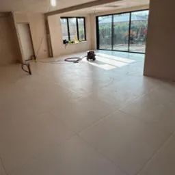 Jasne, kwadratowe płytki podłogowe w dużym, remontowanym pomieszczeniu z dużymi oknami, przez które widać prace ziemne na zewnątrz. Na podłodze stoi czarny odkurzacz przemysłowy. Widoczna...