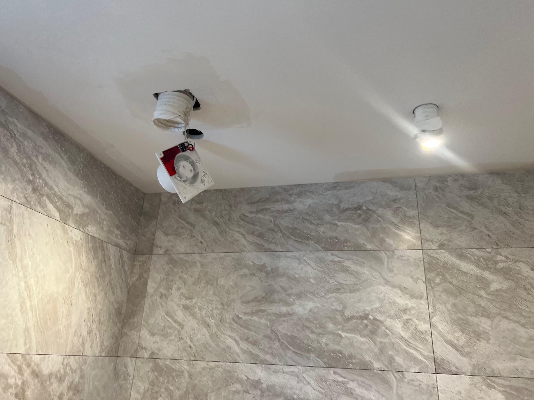 Łazienka w trakcie prac wykończeniowych: marmurowe płytki na ścianie, sufit z otworami na oświetlenie, jeden otwór z podłączoną lampą, drugi z zabezpieczeniem i instrukcją.