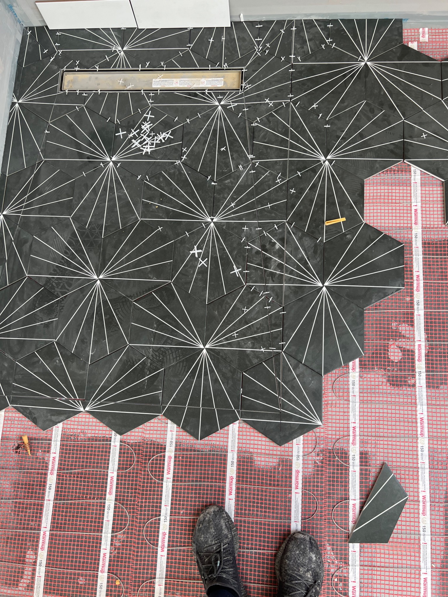 Układanie heksagonalnych, czarnych płytek z białym wzorem na ogrzewaniu podłogowym w łazience w Wągrowcu, widoczny odpływ liniowy i krzyżyki dystansowe.