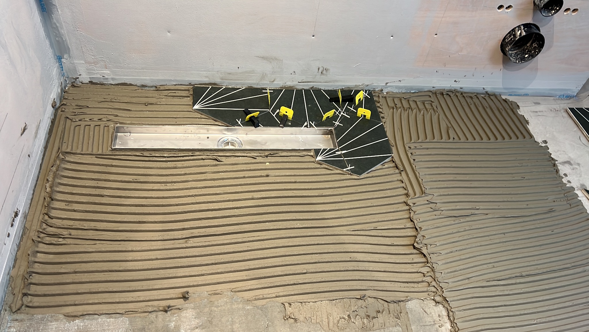 Przygotowanie podłoża pod płytki w łazience z odpływem liniowym, widoczne fragmenty ułożonych płytek z białym wzorem oraz żółte kliny dystansowe.