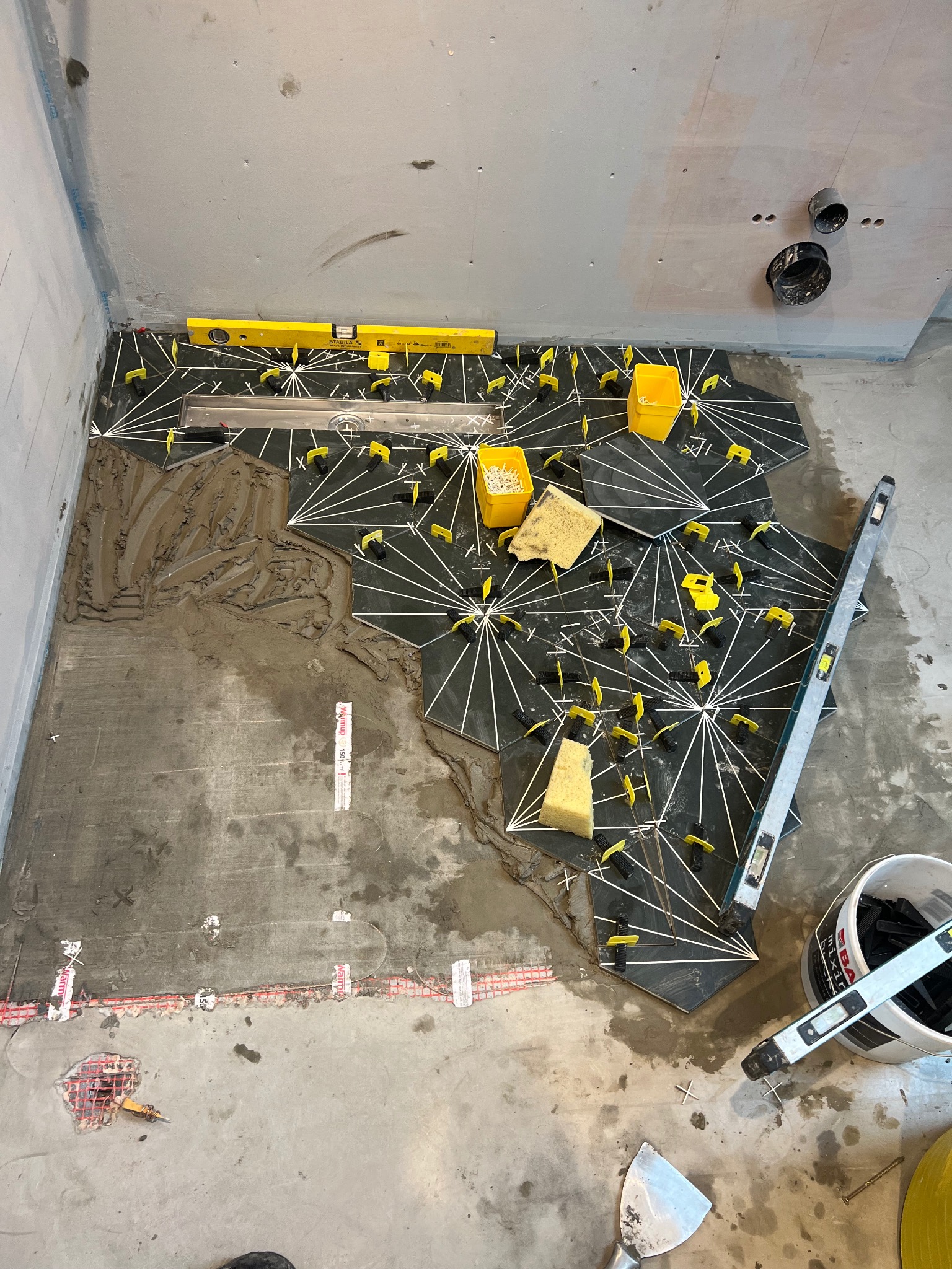 Podłoga w trakcie układania czarno-białych płytek heksagonalnych, widoczne poziomice, krzyżyki dystansowe i resztki zaprawy klejowej, w tle przygotowana ściana pod wykończenie.