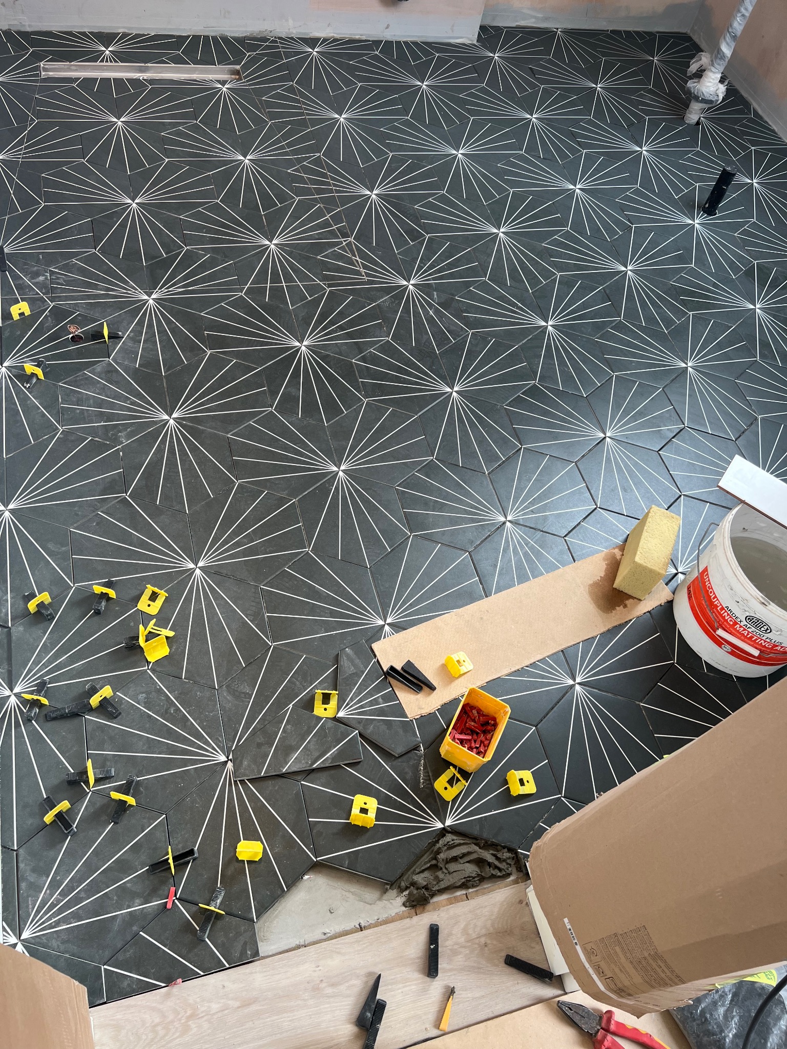 Układanie sześciokątnych, grafitowych płytek podłogowych z białym, geometrycznym wzorem. Widoczne żółte i czarne klipsy poziomujące, wiaderko z klejem, gąbka i fragment podłogi przed położeniem...