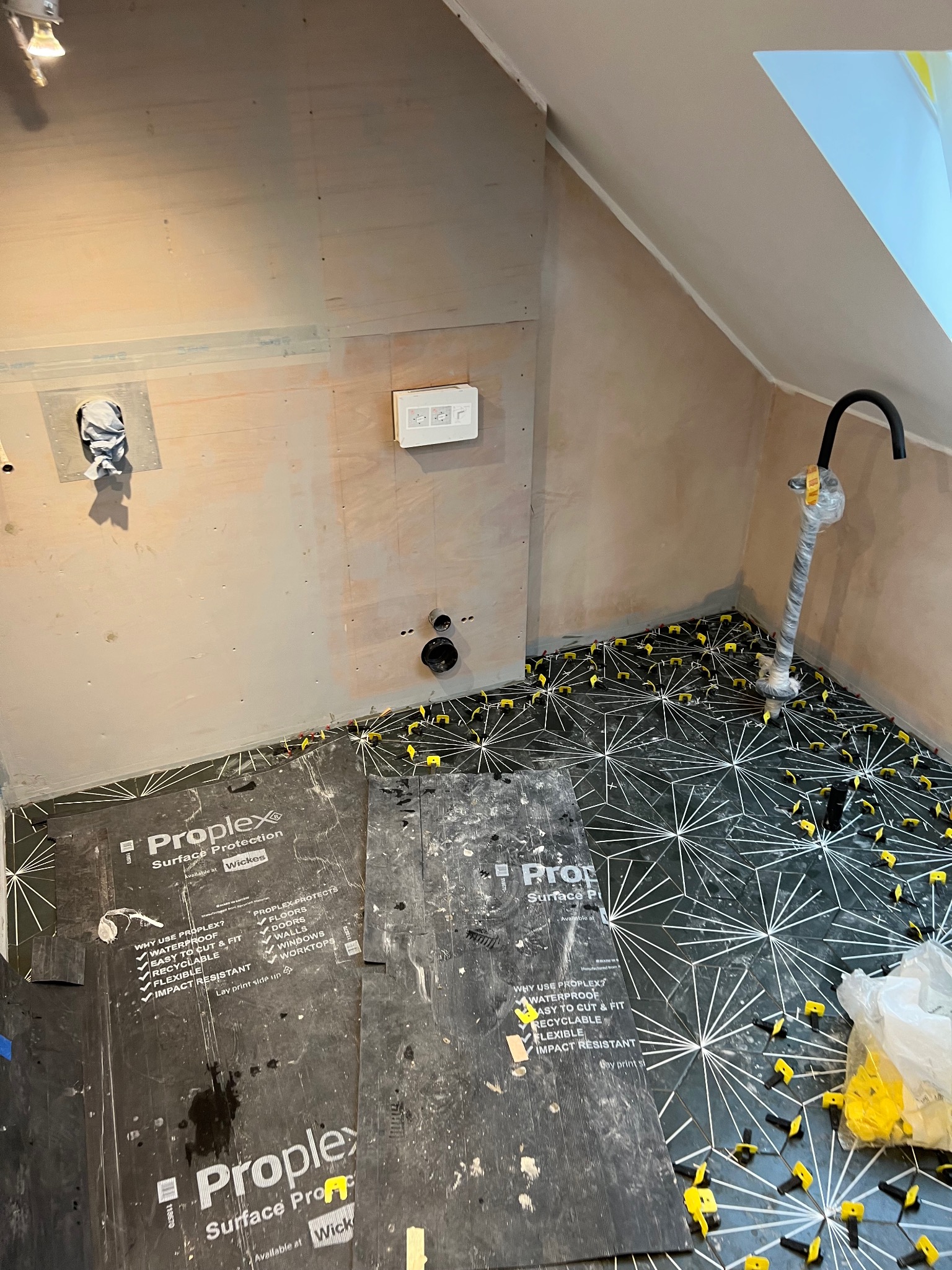 Układanie czarno-białej mozaiki podłogowej w łazience na poddaszu, z użyciem klinów poziomujących i arkuszy ochronnych Proplex Surface Protection.