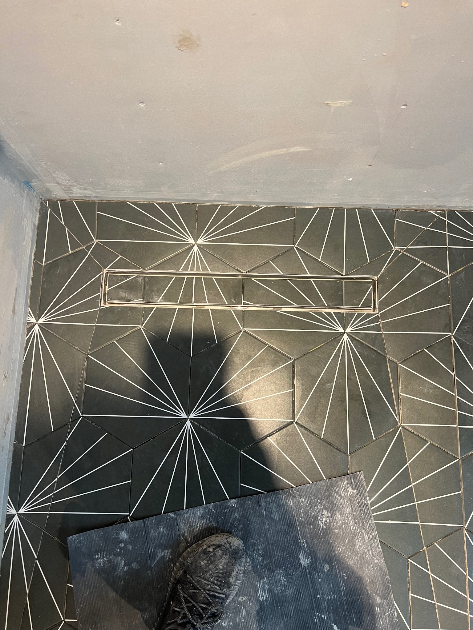 Czarno-biała, geometryczna posadzka w łazience z odpływem liniowym, częściowo zakryta brudną deską i butem roboczym.