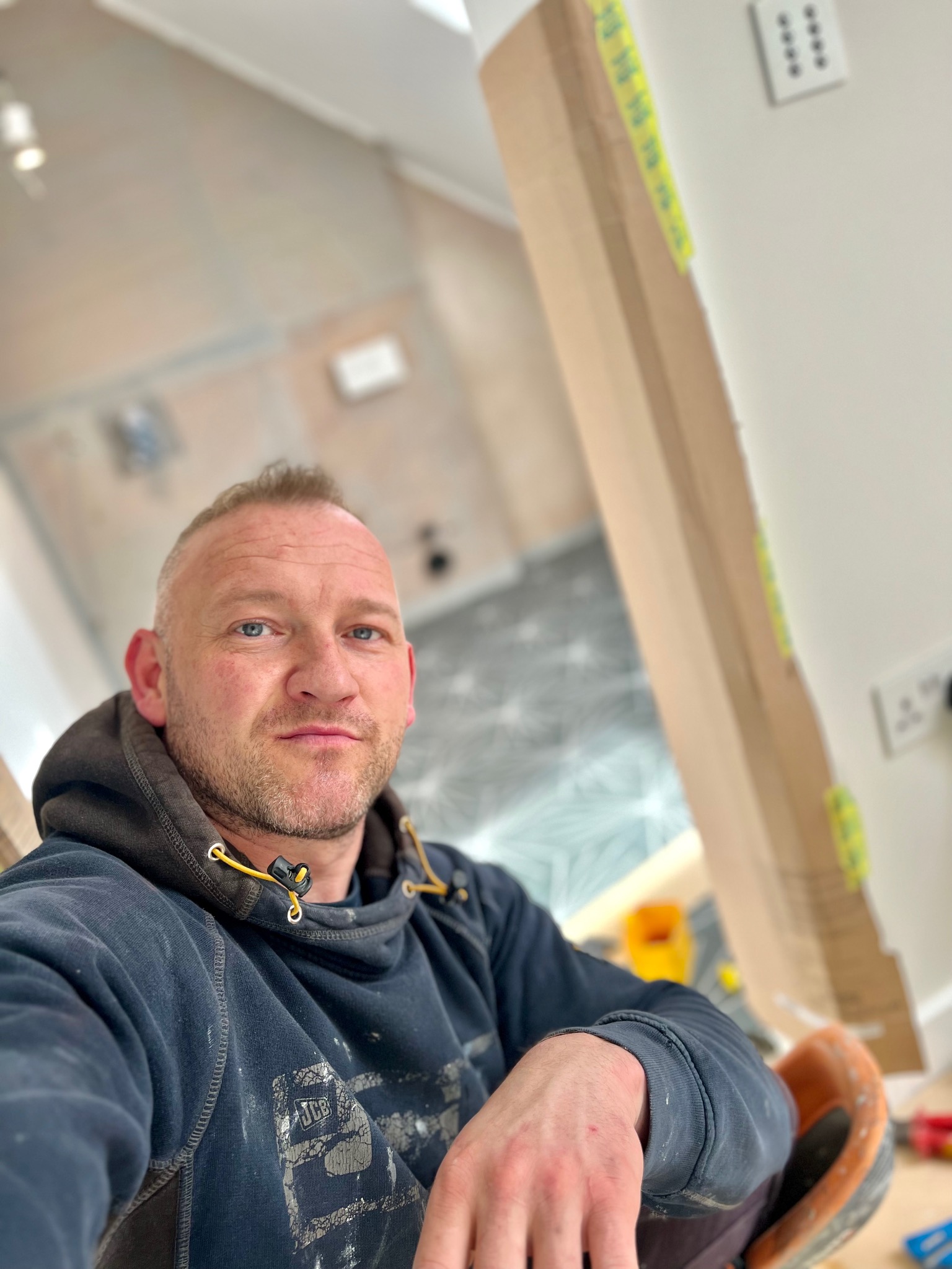 Autoportret mężczyzny w bluzie z kapturem na tle remontowanej łazienki z widoczną podłogą w geometryczne wzory i zabezpieczonymi kartonem framugami drzwi.