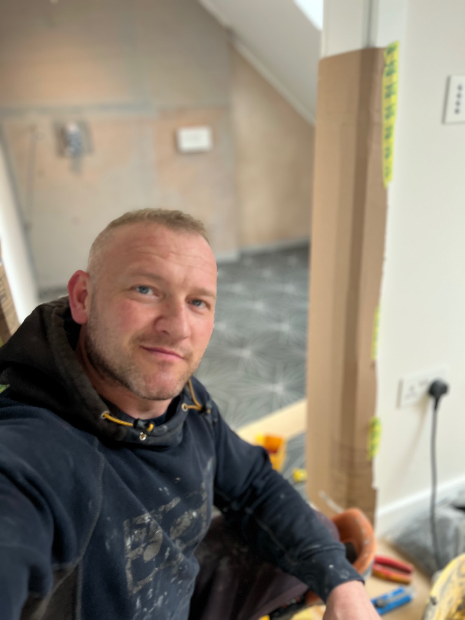 Autoportret rzemieślnika w bluzie roboczej z plamami farby, na pierwszym planie narzędzia, w tle świeżo ułożona podłoga z geometrycznym wzorem w szarościach i fragment ściany w trakcie prac...