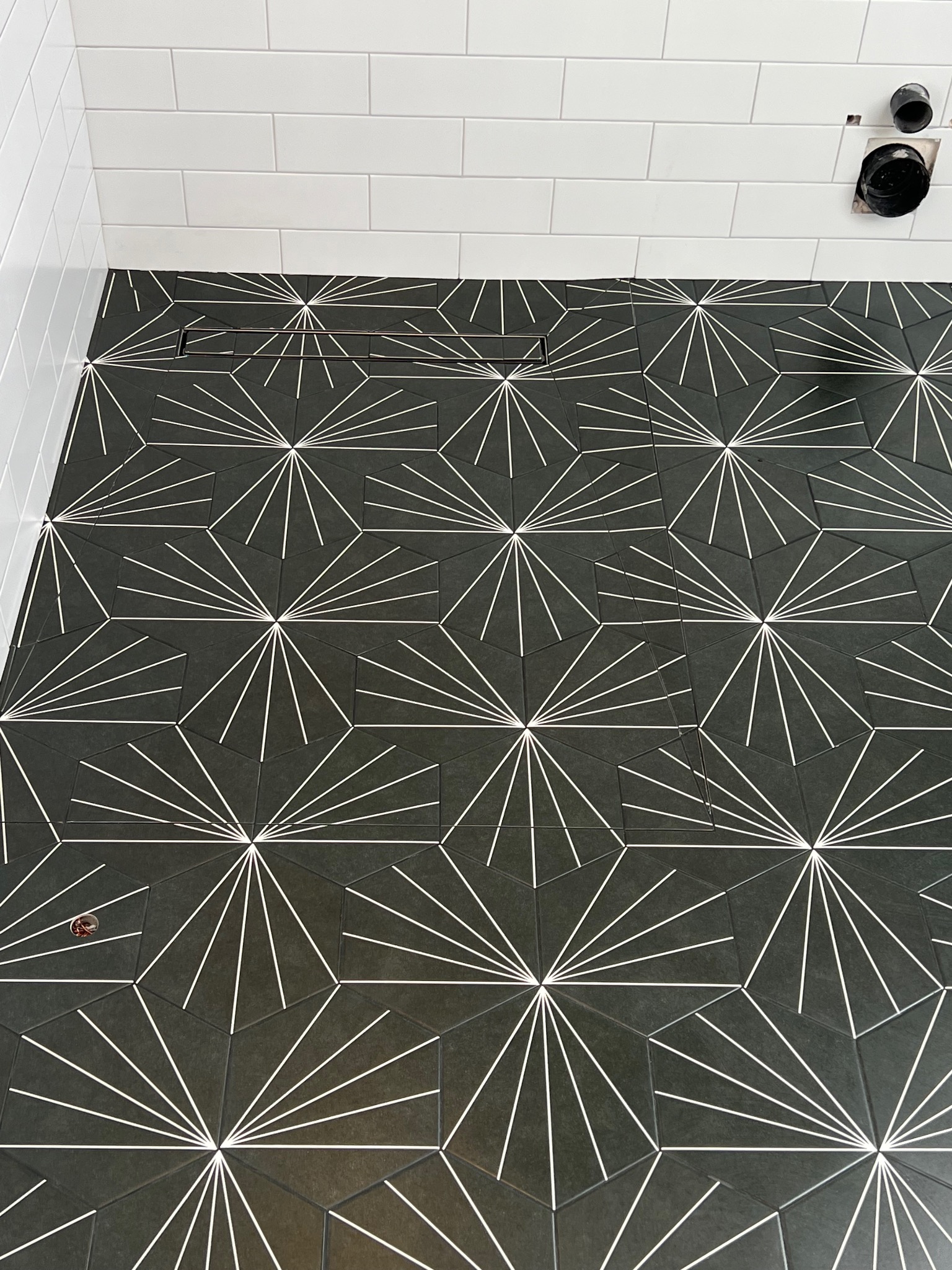 Czarno-biała posadzka z geometrycznym wzorem w łazience, widoczny odpływ liniowy i fragment ściany z białymi płytkami, niedokończone przyłącza rur na ścianie.