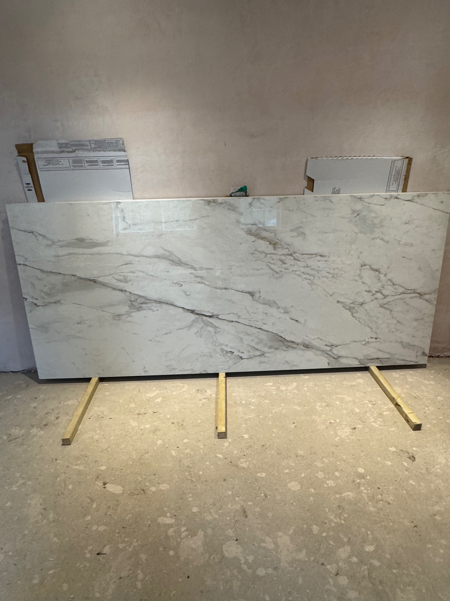 Duża płyta z wzorem marmuru, oparta na trzech drewnianych listwach w surowym wnętrzu z szarą ścianą i podłogą przed montażem.