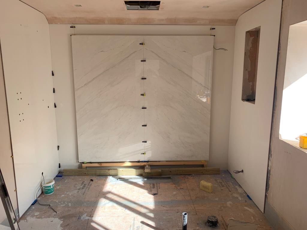 Montaż dwóch dużych płyt marmurowych na ścianie w trakcie remontu łazienki, z użyciem poziomicy, drewnianych podpór i klipsów poziomujących.