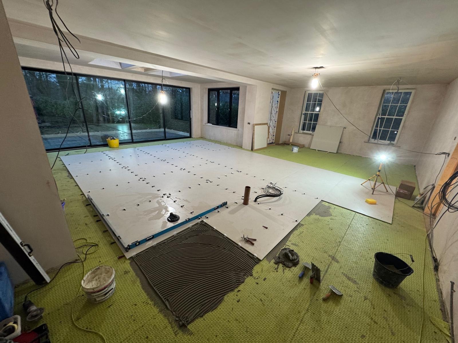 Wnętrze w trakcie układania jasnych, wielkoformatowych płytek podłogowych z użyciem poziomicy i krzyżyków dystansowych; widoczny fragment nałożonego kleju i narzędzia glazurnicze.