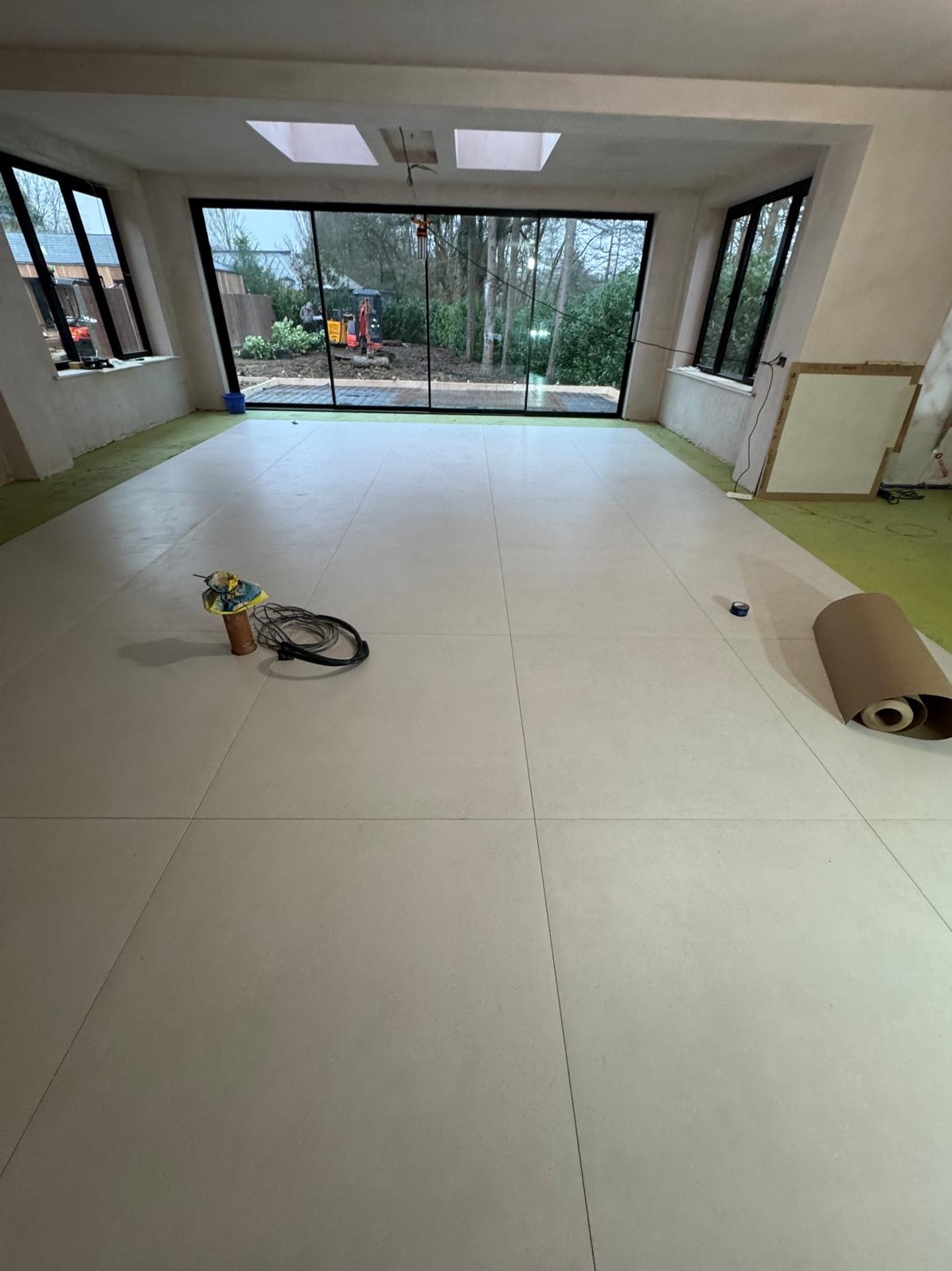 Ułożona jasna podłoga z płytek w przestronnym pomieszczeniu z dużymi oknami z widokiem na zewnątrz, narzędzia i rolka papieru ochronnego na podłodze.