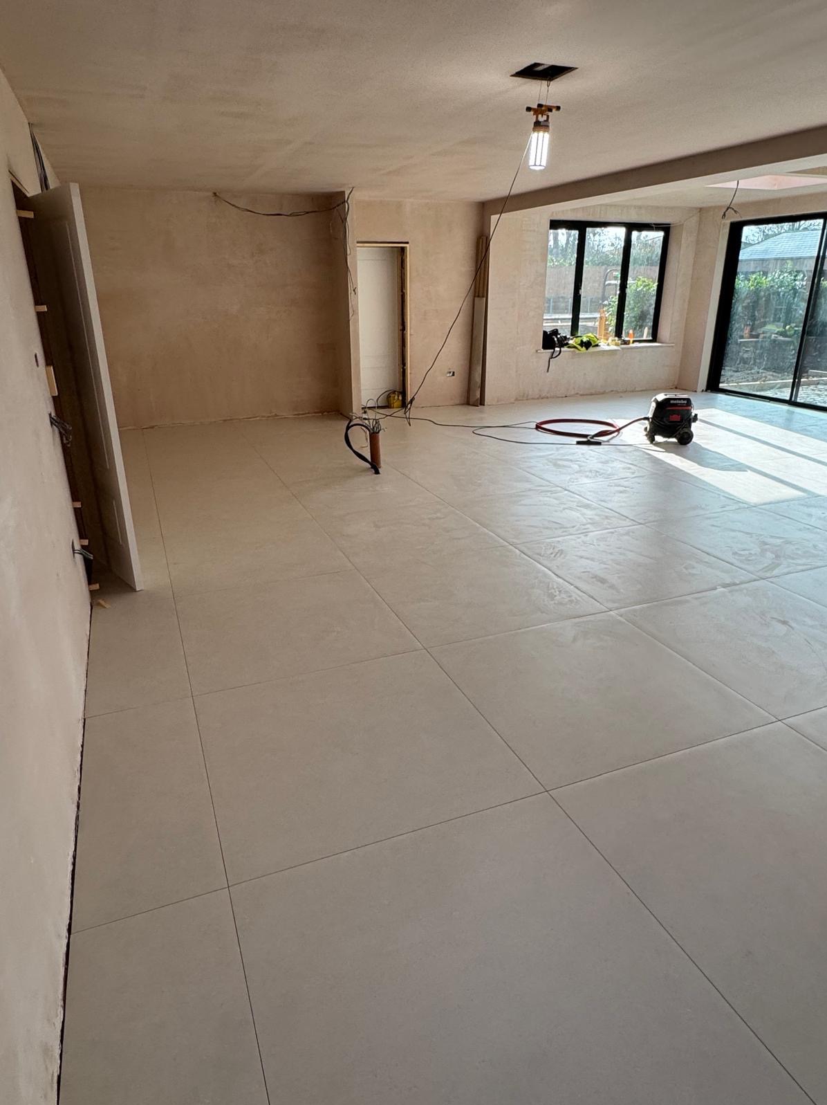 Wnętrze w trakcie remontu z położoną jasną, kwadratową glazurą podłogową, widoczne narzędzia i odkurzacz przemysłowy, ściany przygotowane do malowania.