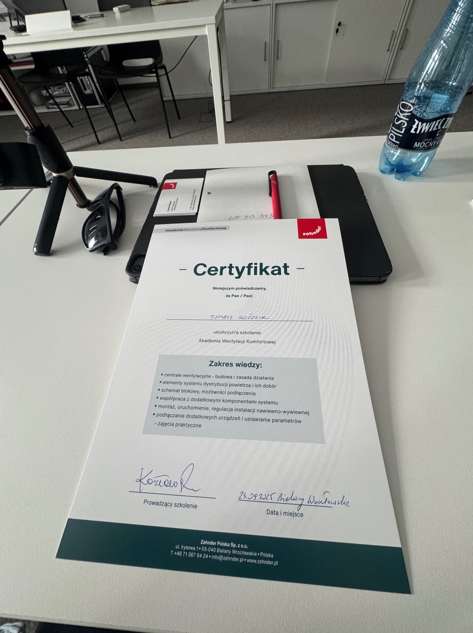 Certyfikat ukończenia szkolenia z wentylacji komfortowej, wystawiony przez Akademię Wentylacji Komfortowej, leżący na biurku obok długopisu i butelki wody.