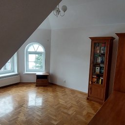 Piotr Wuttke I Andrzej Lotka - Jasne wnętrze poddasza z parkietem w jodełkę, oknami w kształcie łuku i drewnianymi meblami, widok z narożnika pokoju.